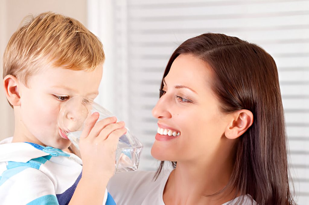 A smiling woman helps a young boy drink water from a clear glass, promoting healthy hydration habits.